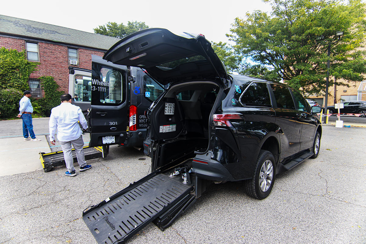 A person standing near an ambulette while another person stands farther away; the ambulette’s back door is open.