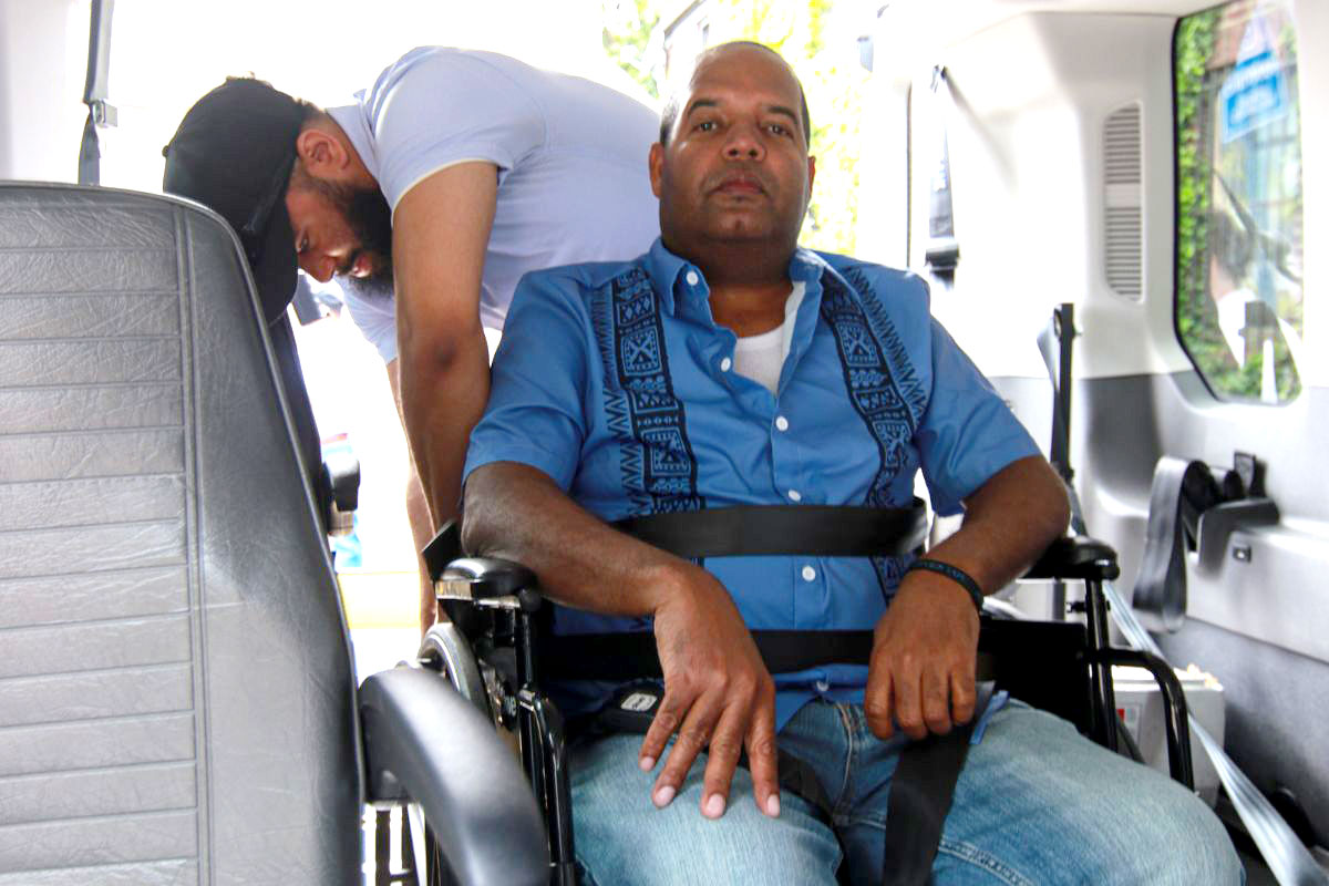 An attendant helps a person with a disability on a wheelchair fasten their seat belt inside an ambulette.
